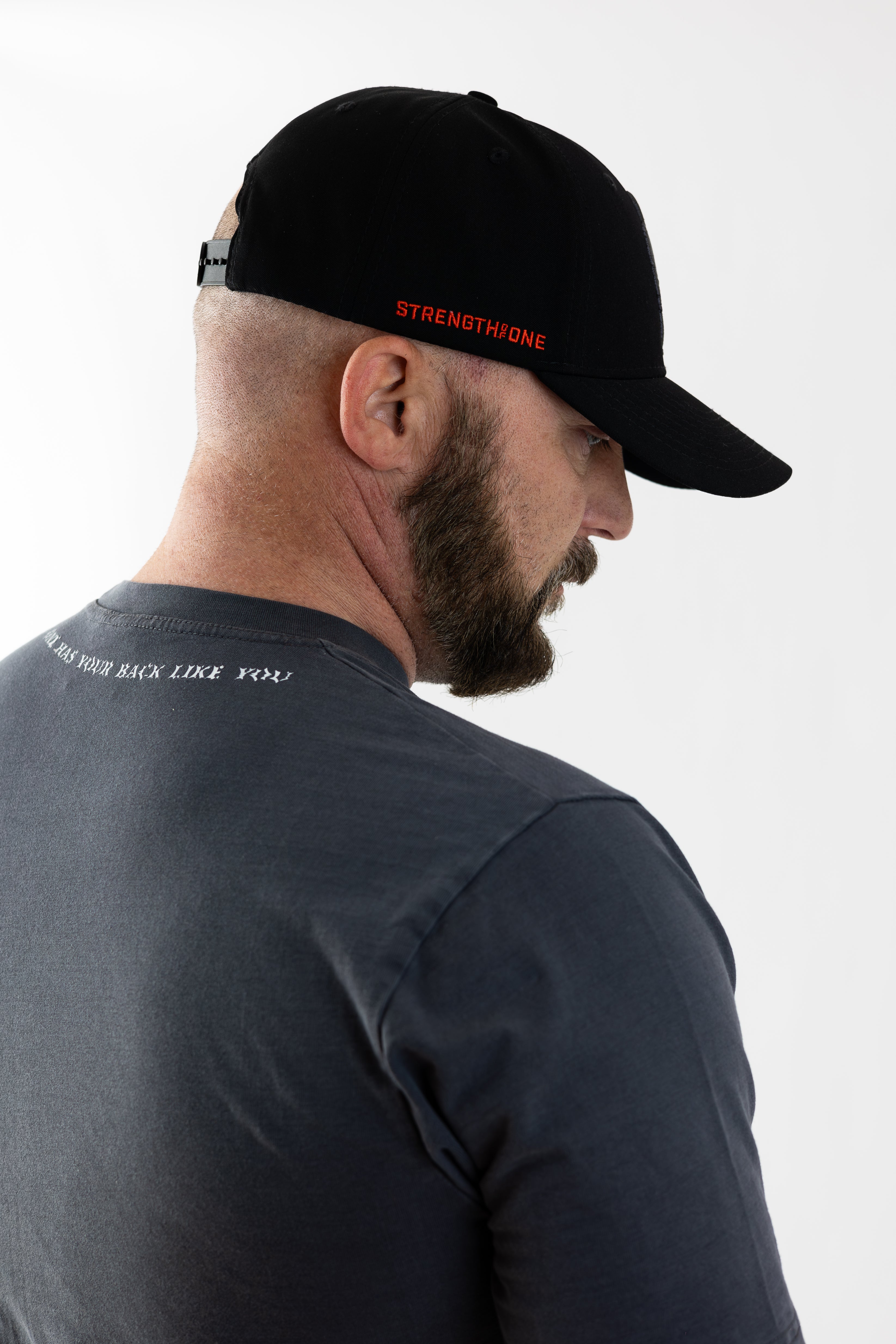 Man in a black Strength of One cap and gray t-shirt viewed from the side against a white background.
