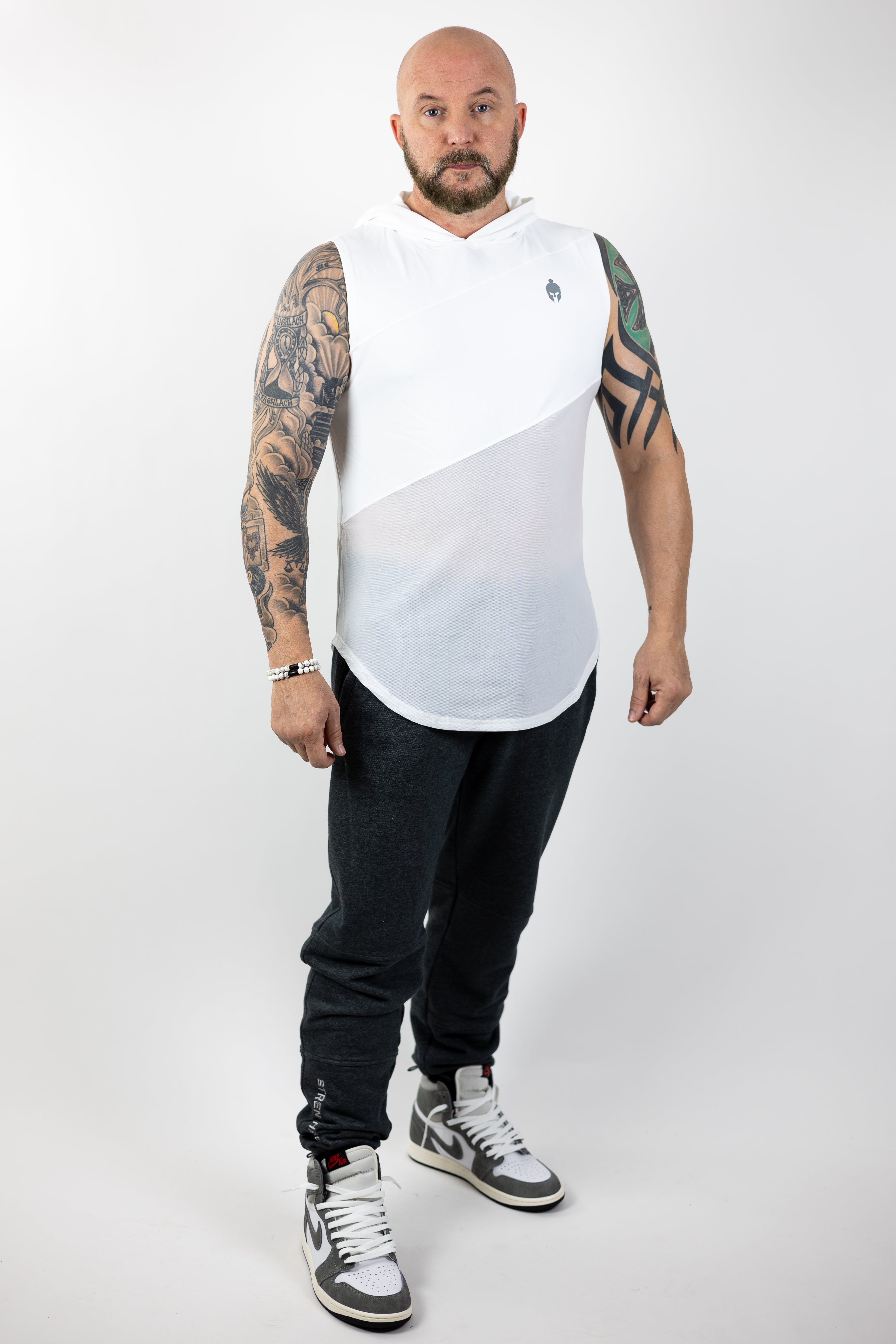 A man with tattoos on his arms stands confidently in a white Strength of One shirt and gray sweatpants, wearing high-top sneakers