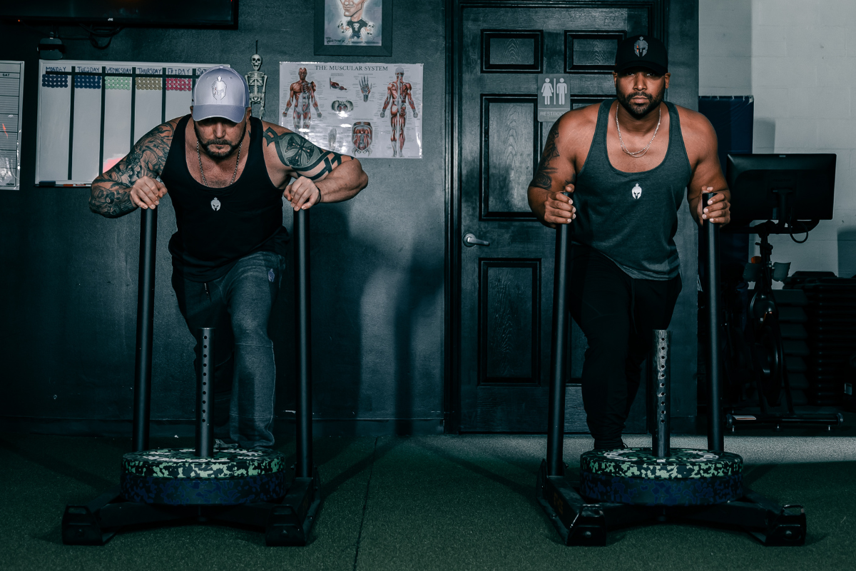 Two men pushing weighted workout sleds wearing Strength of One gym apparel.