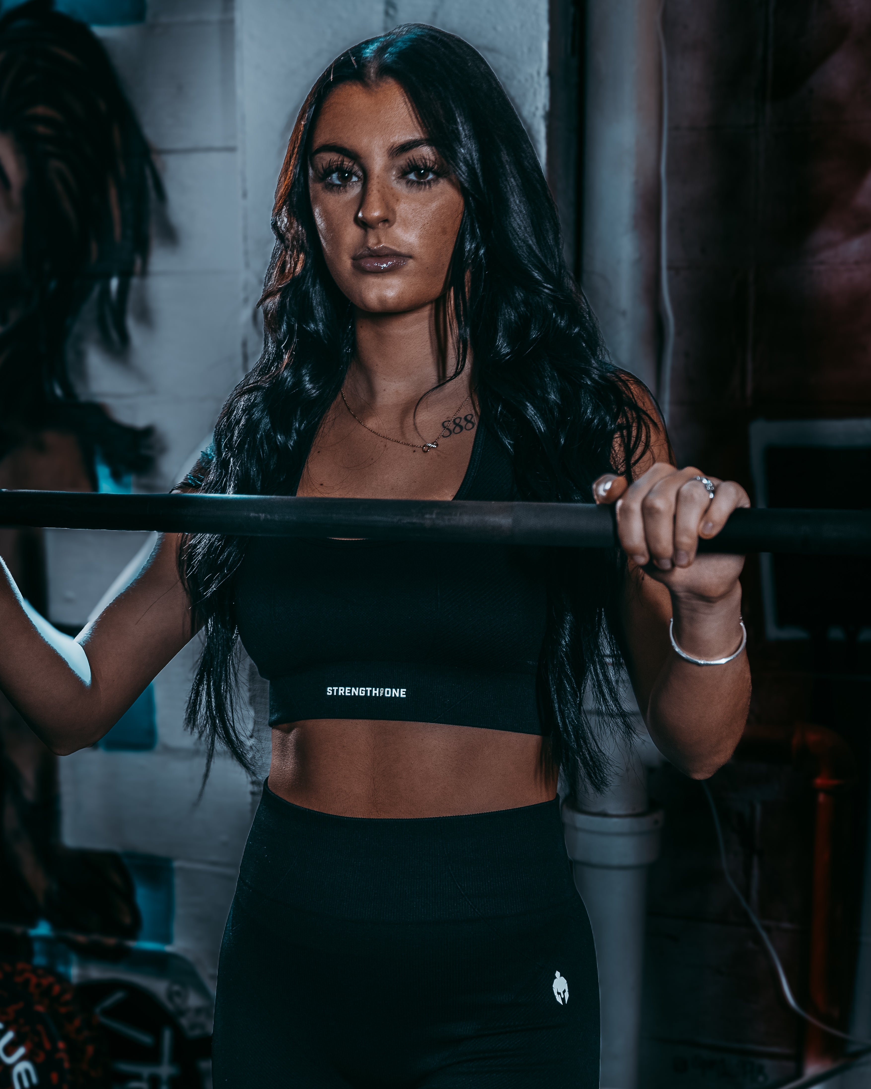 Woman wearing Strength of One sports bra and leggings set gripping squat rack barbell