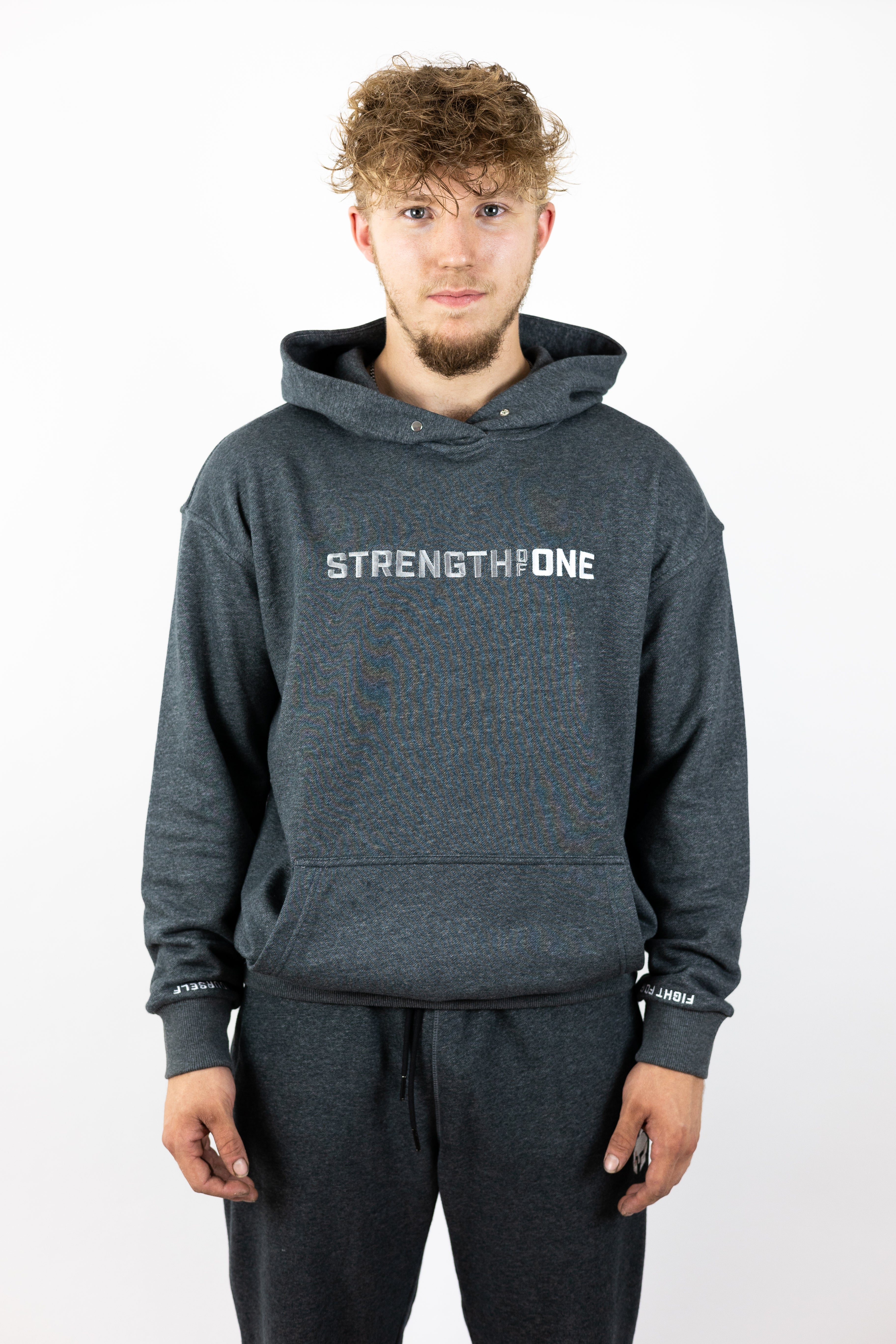 Man standing tall with hands by side and wearing gray hoodie with words Strength of One across chest