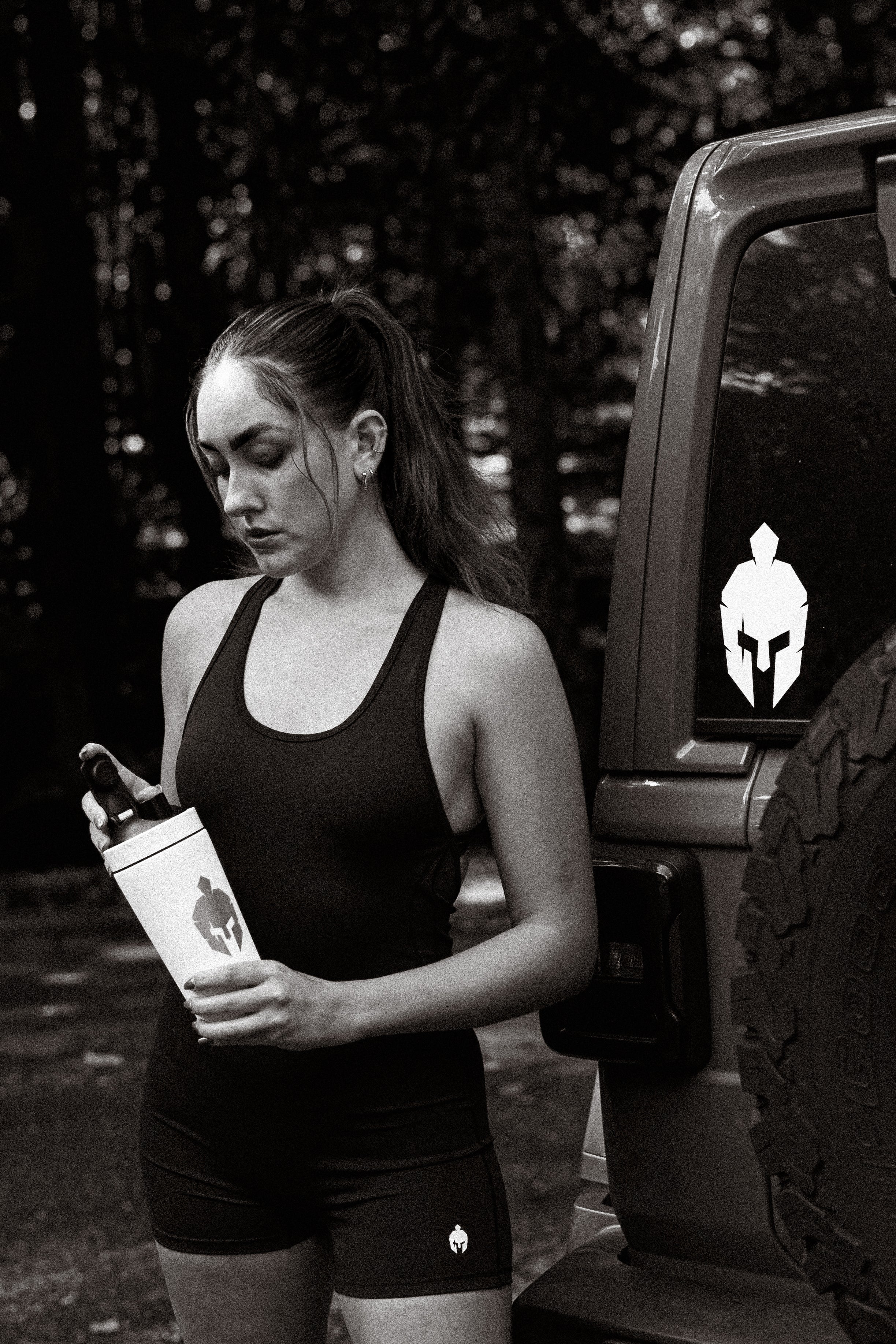 A woman in Strength of One athletic wear checking her shaker bottle next to a vehicle with a warrior helmet decal
