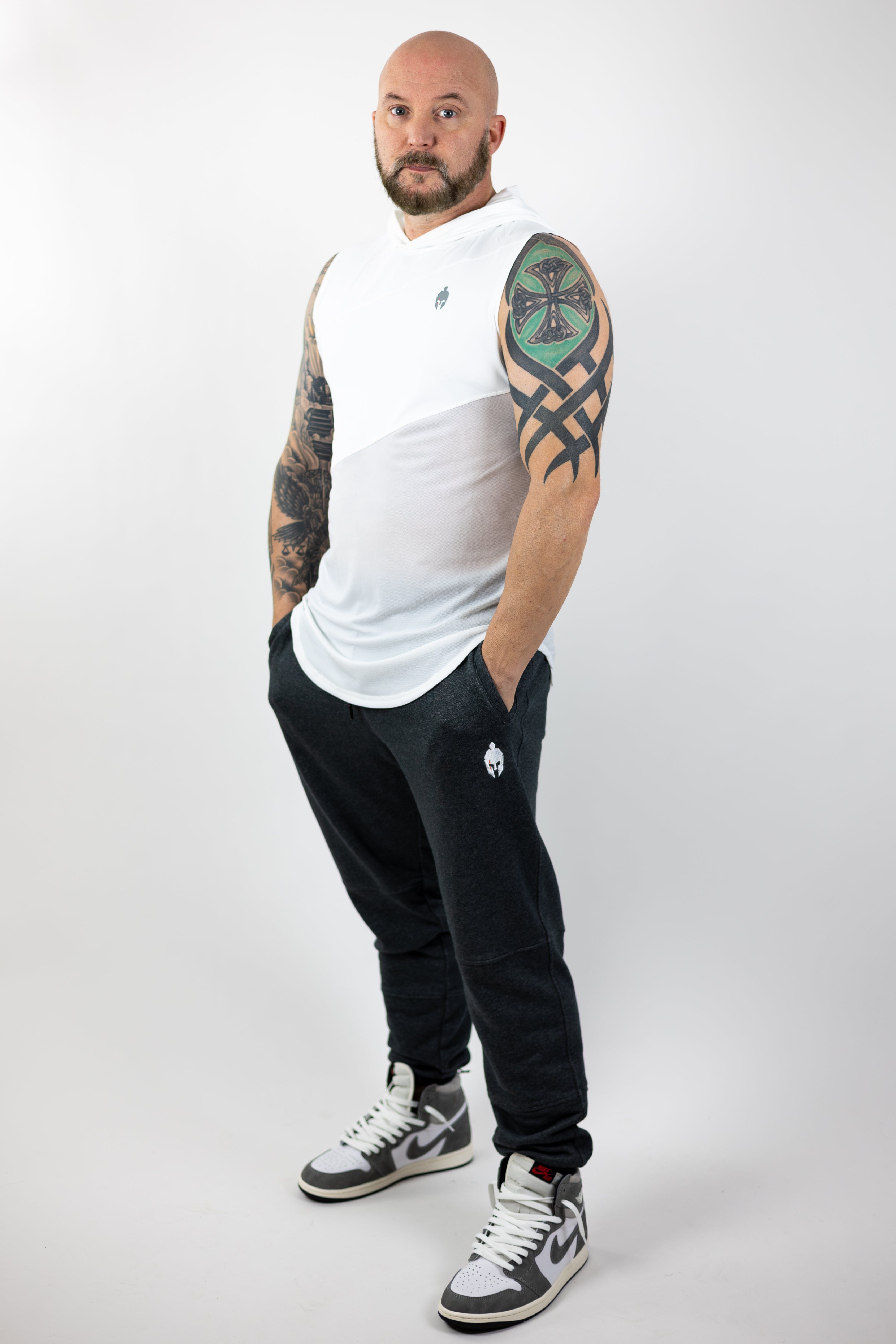 A man with tattoos on his arms stands confidently in a white Strength of One shirt and gray sweatpants, wearing high-top sneakers