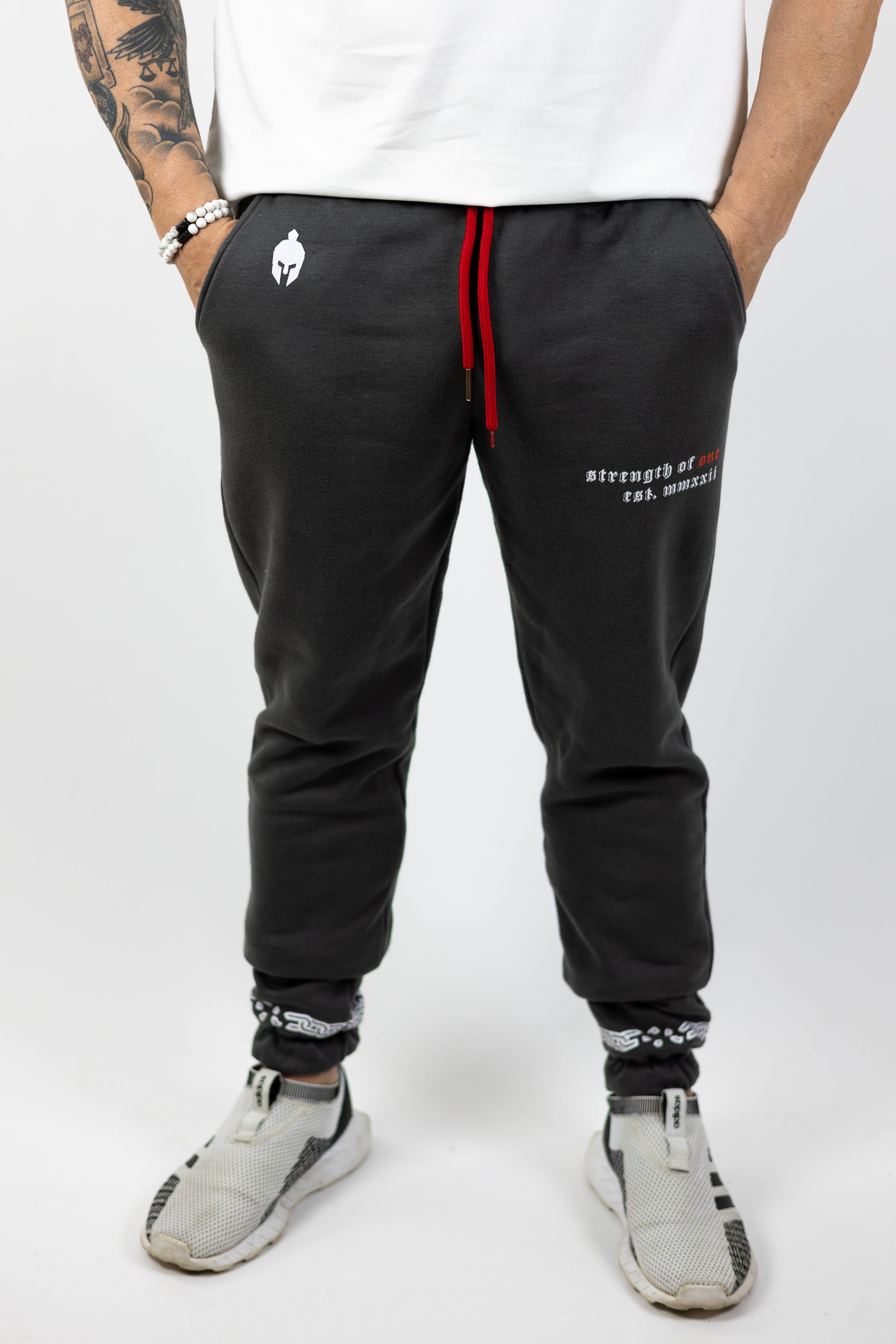 man wearing gray Strength of One gym sweatpants with warrior helmet logo on top left hip and Strength of One logo on right hip with red drawstrings coming out of pants