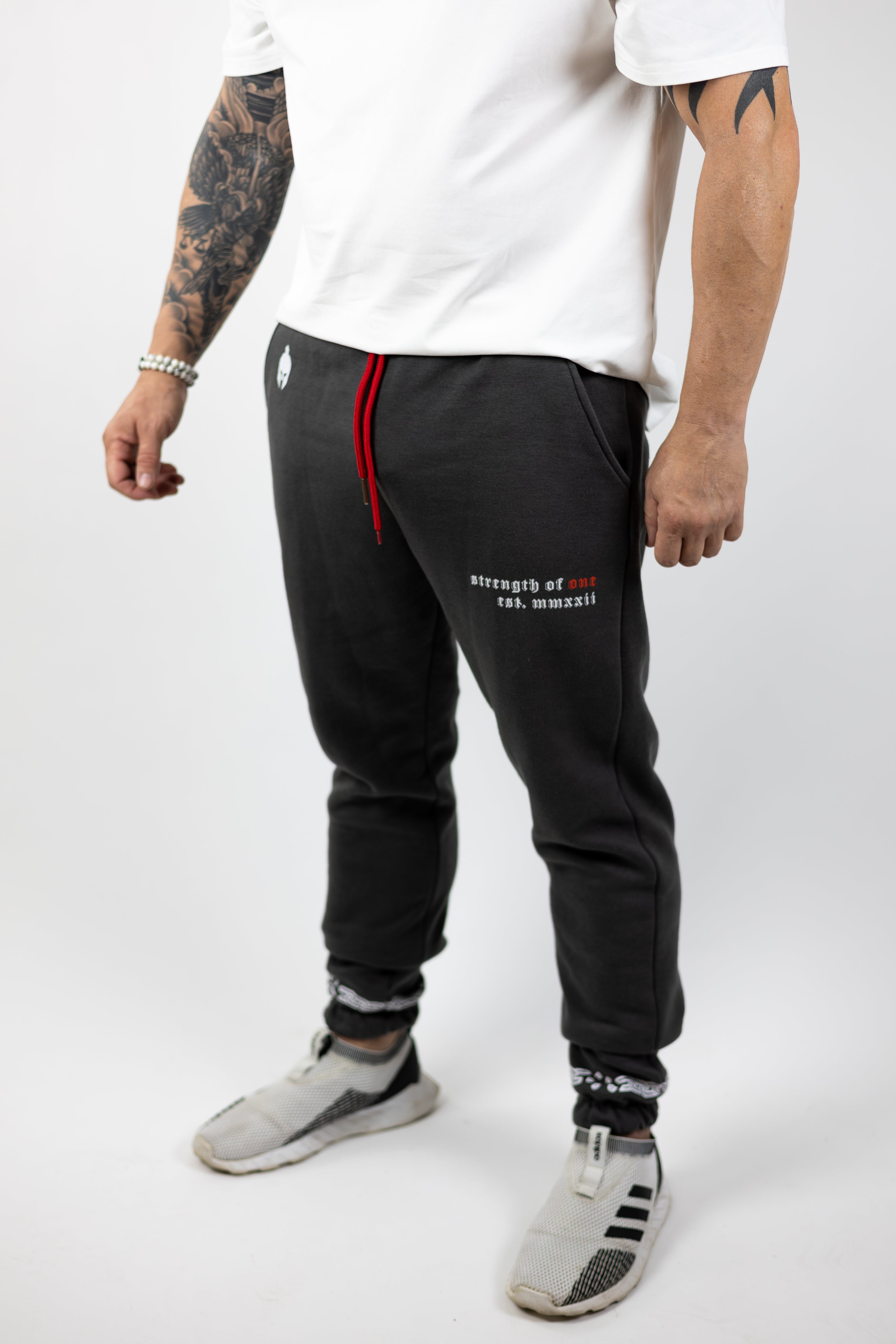 man wearing gray Strength of One gym sweatpants with warrior helmet logo on top left hip and Strength of One logo on right hip with red drawstrings coming out of pants