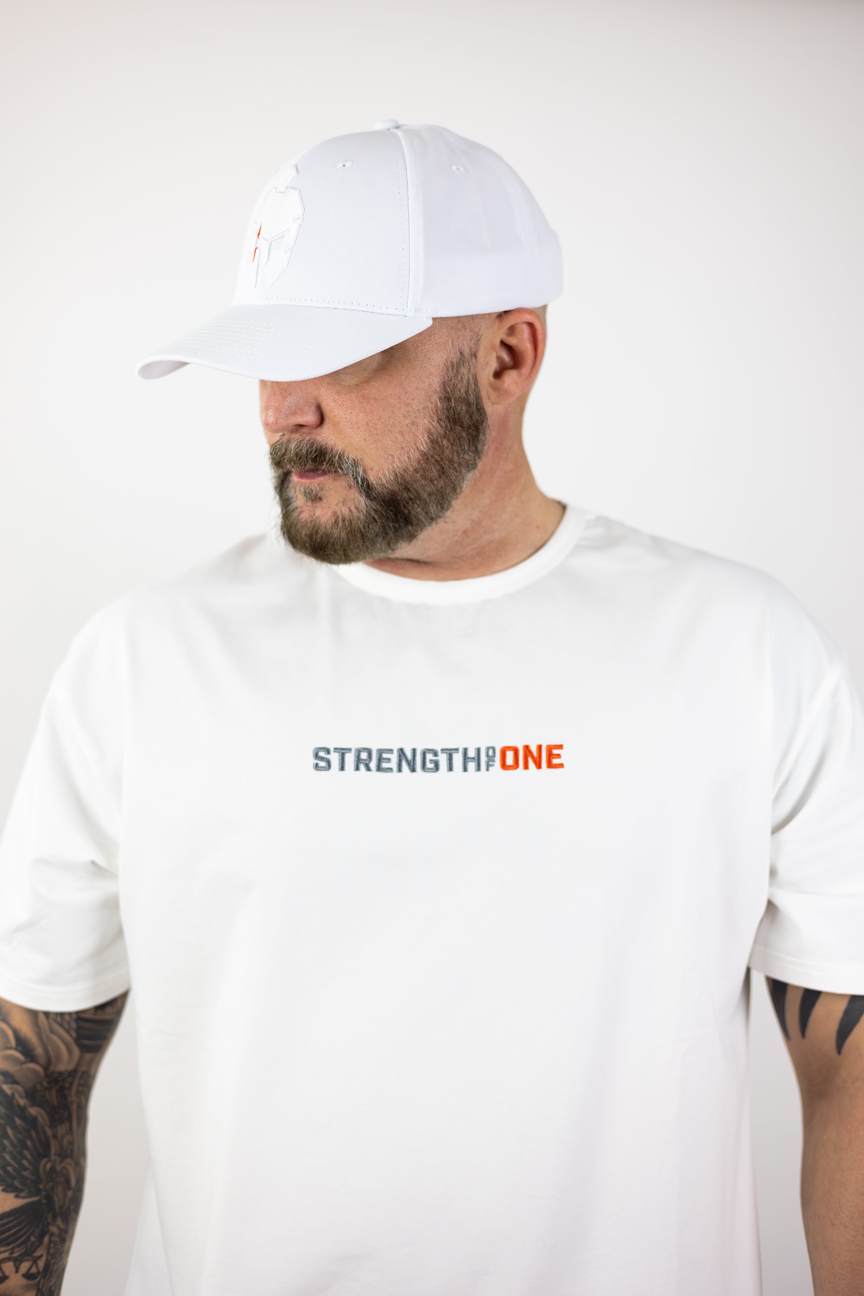 man modeling white Strength of One workout shirt with an embroidered logo reading Strength of One and wearing white baseball cap with spartan warrior helmet logo