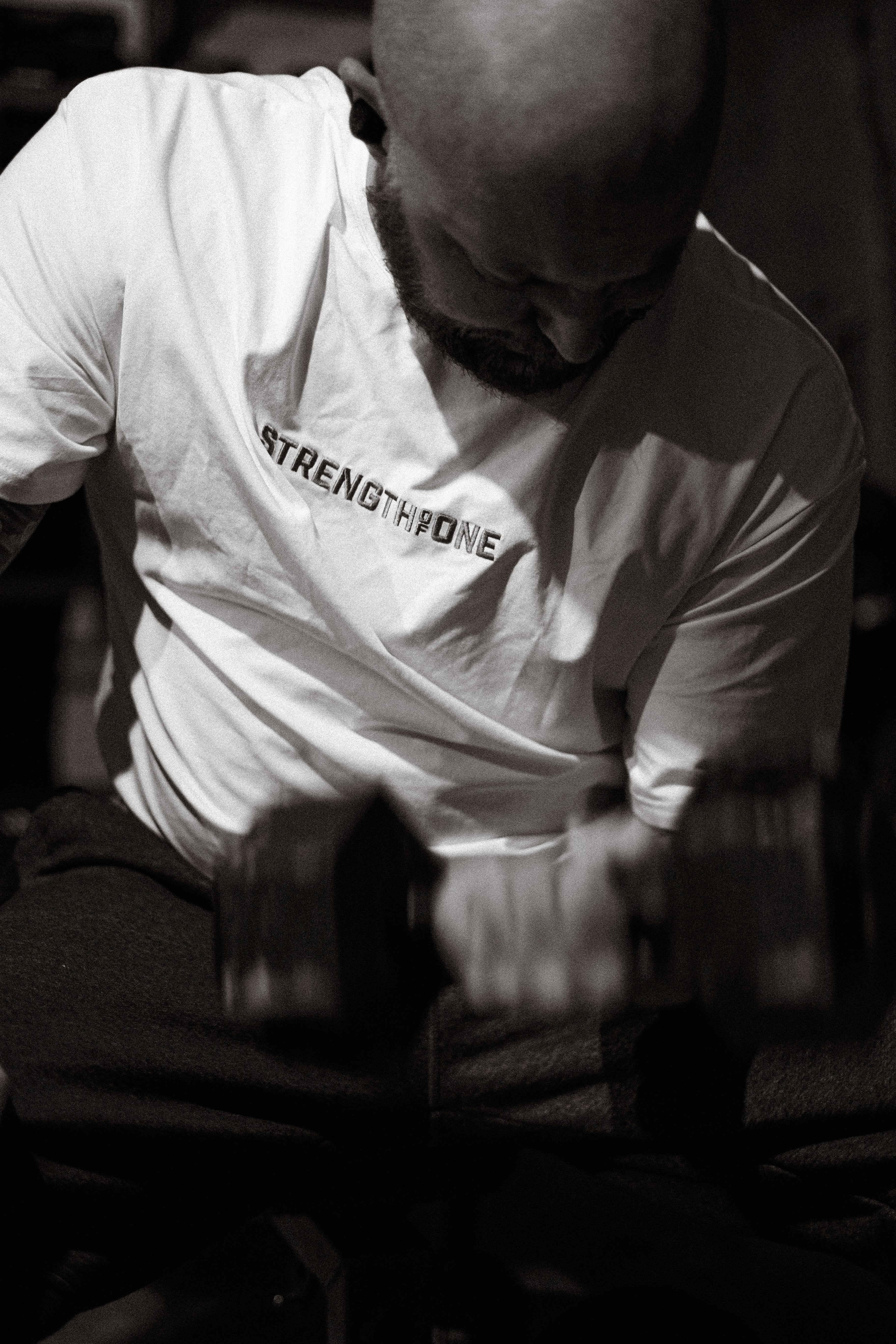 Man in a Strength of One white t-shirt lifting dumbbells in a gym setting.