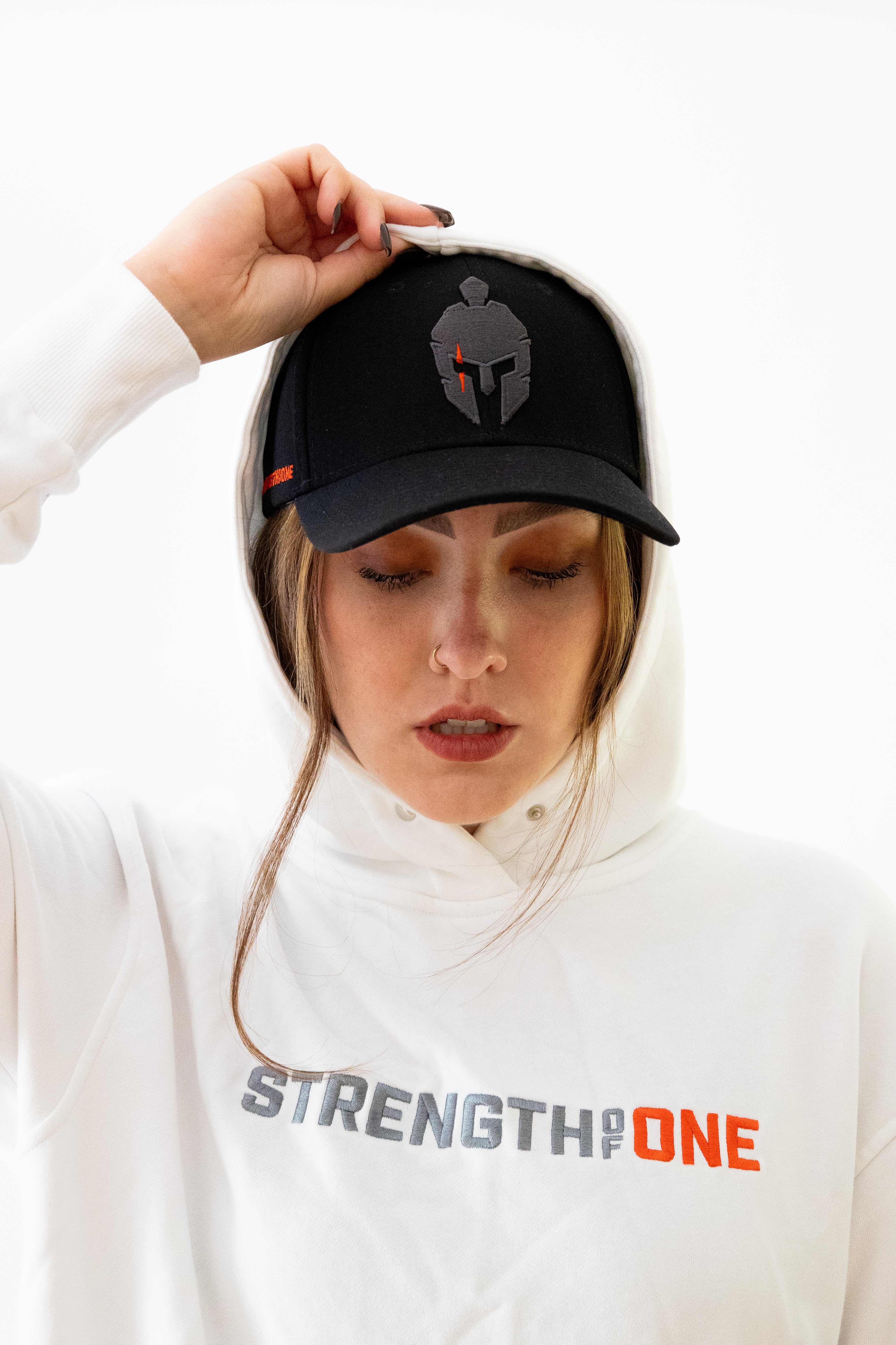 Woman wearing white Strength of One hoodie putting hood over Strength of One cap